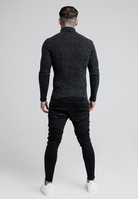 Schwarzes, geripptes Langarmshirt mit hohem Kragen, kombiniert mit schmal geschnittenen schwarzen Jeans und weißen Turnschuhen, von hinten betrachtet.