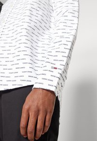 Camicia bianca a maniche lunghe con un motivo ripetuto di testo nero "TOMMY JEANS". Presenta un dettaglio con bottone al colletto e una piccola accentuazione rossa-blu.