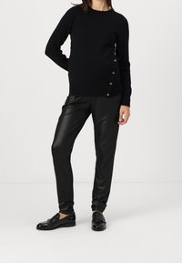 Person wearing a black sweater with side buttons, black faux leather pants, and black loafers standing against a plain white background.