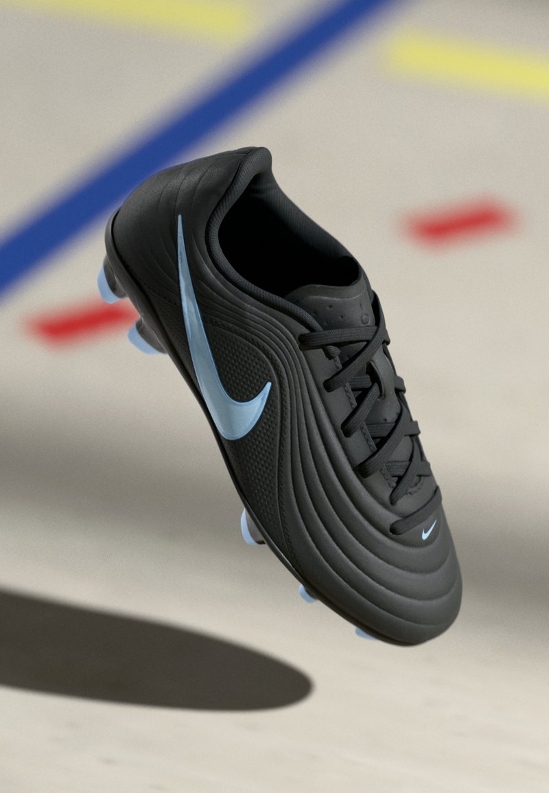 Μαύρο ποδοσφαιρικό παπούτσι Nike με ανοιχτό μπλε swoosh και καρφιά, που αιωρείται πάνω από έναν ανοιχτό δάπεδο με πολύχρωμες γραμμές γηπέδου στο φόντο.