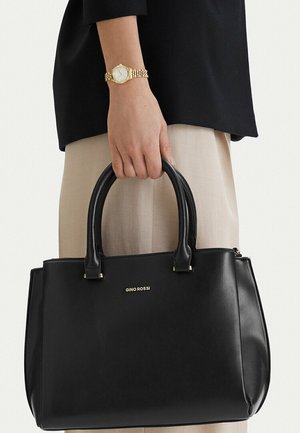 Mano sosteniendo un bolso negro de Gino Rossi con asas dobles, llevando un reloj de pulsera dorado, una blusa negra y pantalones beige.