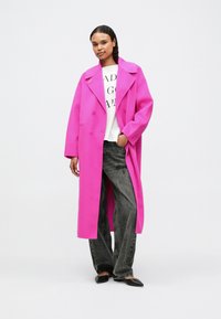 Sieviete stāv ar vienu roku kabatā, ģērbusies spilgti rozā garā mētelī, baltā grafiskā T-kreklā, brīvās melnās džinsās un melnās norādītajās kurpēs.