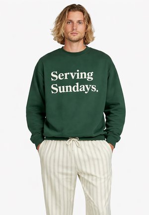 Hombre con cabello rubio de longitud media que lleva una sudadera verde oscuro con el texto "Serving Sundays" y pantalones con cordón de rayas claras.