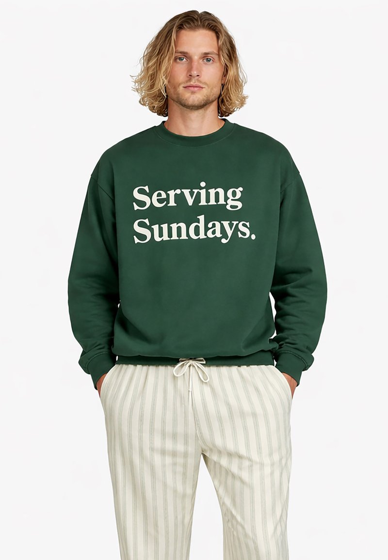 Homme aux cheveux blonds mi-longs portant un sweat-shirt vert foncé avec le texte "Serving Sundays" et un pantalon à cordon de serrage rayé clair.