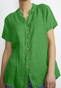Green linen short-sleeve shirt with a mandarin collar, feature buttons in the front, and subtle pleats at the shoulders. Smooth texture.