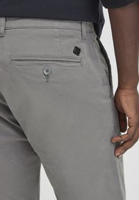 Gray cotton trousers featuring a back pocket with a button closure and a black accent tag near the waistband. Smooth texture and tailored fit.