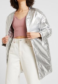 Srebrna puchowa kurtka o błyszczącej, pikowanej strukturze. Posiada opadające ramiona i luźny krój, noszona na różowej bluzce w paski oraz białych dżinsach.