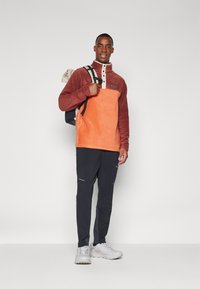 Fleece-pullover i orange och burgundy med hög krage, vit knapprad, parat med mörka byxor och vita sneakers.