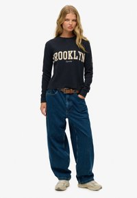 Marineblauw longsleeve shirt met "BROOKLYN" graphic; gecombineerd met losse blauwe jeans en lichtgekleurde sneakers. Katoenen materiaal, casual pasvorm.