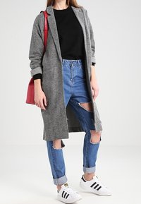 Grijze visgraat lange jas over een zwart shirt, gecombineerd met versleten blauwe jeans. Witte sneakers met zwarte strepen, rode schoudertas.