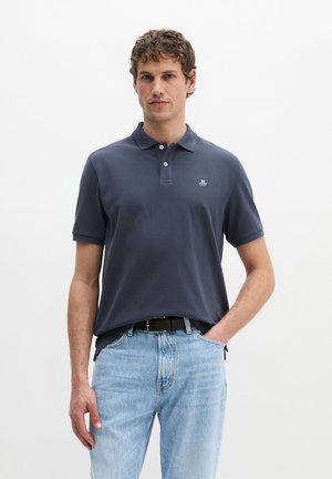 Man met krullend haar, gekleed in een marineblauw poloshirt en lichtblauwe jeans, staat met één hand in de zak tegen een effen achtergrond.