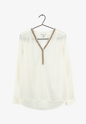 Blouse blanche à manches longues avec un col en V orné de perles dorées et argentées, accrochée à un cintre noir.