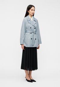 Ανοιχτό μπλε καπαρντίνα με διπλό πέτο, μαύρα κουμπιά και ζώνη στη μέση, συνδυασμένη με πλισέ μαύρη φούστα και μαύρες ψηλοτάκουνες γόβες.