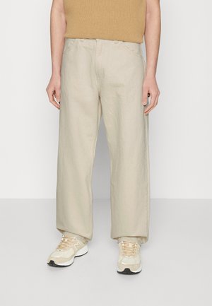 Beige weit geschnittene Hose aus leichtem Stoff mit glatter Textur, ausgestattet mit vorderen Taschen und Gürtelschlaufen, kombiniert mit beigen Sneakers.