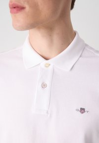 Hvit polo skjorte med klassisk krage, med en liten brodert logo og to knapper, laget av teksturert bomullsmateriale.