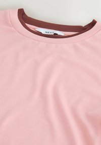 Deux t-shirts à col rond pliés, un rose posé sur un marron, avec une étiquette blanche visible affichant le texte « NEXT » sur un fond uni.