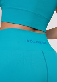 Completo sportivo turchese con texture a coste. Include un top corto e leggings a vita alta, con il logo "OCEANSAPART" in blu.