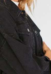 Person, die eine schwarze Jeansjacke mit Metallknöpfen und Brusttaschen trägt, mit über dem Oberkörper gekreuzten Armen vor einem schlichten Hintergrund.