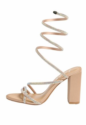 Bézs magas sarkú szandál blokk sarokkal és vékony, ékszeres spirálpánttal, amely a bokát körülöleli.