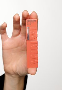 Rossetto liquido corallo in un tubetto arancione rettangolare con testo in rilievo. Il tappo presenta la scritta "Superstay Vinyl Ink" in lettering argentato.