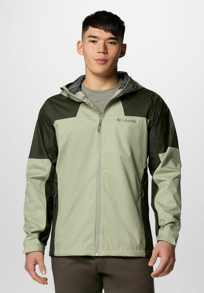 Młody mężczyzna w zielonej i oliwkowej kurtce Columbia z kapturem, stojący na jednolitym jasnym tle, twarzą do przodu.