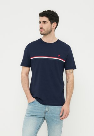 Férfi tengerészkék pólóban, amelyen piros, fehér és szürke vízszintes csíkok vannak a mellkasán, világoskék farmernadrágban, balra néz, kezét a zsebében tartva.