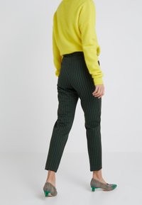 Person som bär en gul tröja, gröna och svarta rutiga byxor samt rutig lågklackade skor med gröna tåhattar, står framför en vit bakgrund.