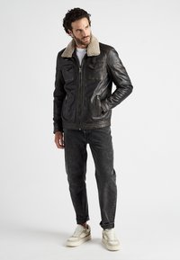 Zwarte leren jas met een beige nepschapenvachtkraag; voorzien van twee borstzakken en zijritszakken; heeft een gestructureerde afwerking; gedragen met een donkere spijkerbroek en sneakers.