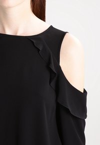 Svart topp med kall axel-design, strukturerad tyg och volangdetaljer längs halsringningen och axelsömmen.