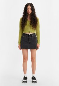 Groene donzige cropped cardigan met knoopdetails, gecombineerd met een zwarte denim mini-jurk met een rafelige zoom en zwart-witte sneakers.