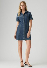Denim shirtjurk, getailleerd design, korte mouwen, knopenfront, twee borstzakken, onafgebroken zoom, donkerblauwe kleur, gecombineerd met zwarte sandalen met bandjes.