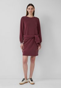 Bordeauxfarbenes langärmliges Kleid mit einer gebundenen Taillenpartie. Aus weichem Stoff gefertigt, hat es einen runden Ausschnitt und kontrastierende Streifen an den Bündchen.