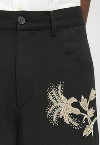 Μαύρο υφασμάτινο παντελόνι με πολύπλοκα χρυσά κεντήματα ανθισμένων σχεδίων, με κουμπί και πλευρικές τσέπες. Υφή υλικού.