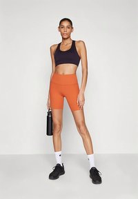 Ženska v črnem športnem modrčku in oranžnih kolesarskih kratkih hlačah drži črno steklenico za vodo, nosi bele nogavice in črne superge, stoji ob ozadju enobarvne stene.