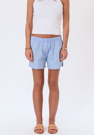 Kvinde iført lyseblå elastiske shorts med talje, hvid ærmeløs top, guldbøjler, ur, ringe og beige sandaler med kædedetalje.