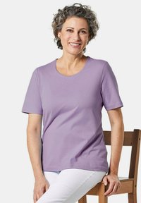 Lila katoenen T-shirt met korte mouwen en een ronde hals, gecombineerd met witte broek. Model zit op een houten stoel.