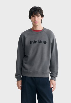 Jeune homme aux cheveux foncés portant un sweat-shirt gris avec le texte "thinking." et un pantalon foncé, debout devant un fond clair uni.