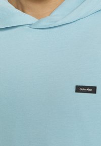 Felpa leggera blu chiaro realizzata in materiale morbido con tasca a canguro. Presenta un piccolo logo nero di Calvin Klein ricamato sul petto.