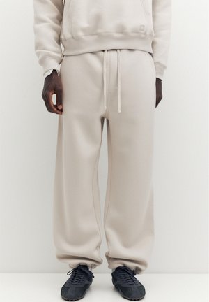Homme portant un pantalon de survêtement beige et un sweat assorti, baskets noires, debout devant un fond uni clair.