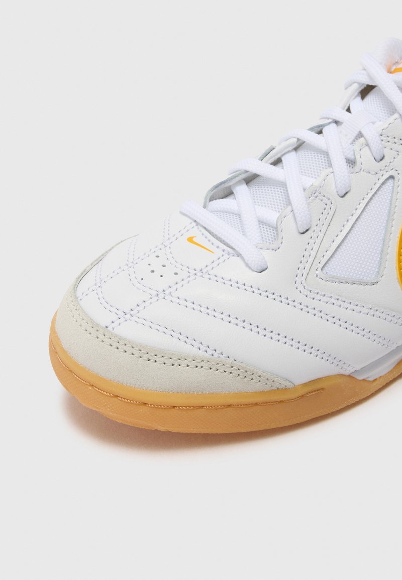 Zapatilla deportiva blanca fabricada con materiales de ante y cuero sintético, con acentos naranjas, diseño texturizado y una suela de goma. Los cordones son blancos.
