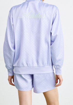 Personne portant une veste Adidas Originals violet clair et un short assorti, vue de dos sur un fond uni.