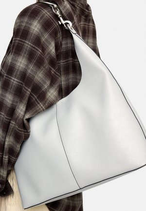 Biała skórzana torba na ramię z czarnym obszyciem, noszona na ramieniu osoby ubrane w brązową koszulę w kratę.