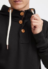 Felpa nera con cappuccio caratterizzata da una zip a quarto, bottoni di legno decorativi e un tessuto testurizzato con lacci color crema.