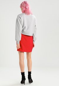 Osoba s růžovými vlasy, oblečená v šedém dlouhém rukávu, červené sukni a černých vysokých podpatcích, stojící zády k bílému pozadí.