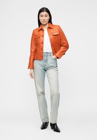 Γυναίκα που στέκεται φορώντας πορτοκαλί δερμάτινο μπουφάν, λευκό πουκάμισο, ανοιχτό μπλε τζιν και μαύρα μποτάκια αστραγάλου, μπροστά σε απλό φόντο.