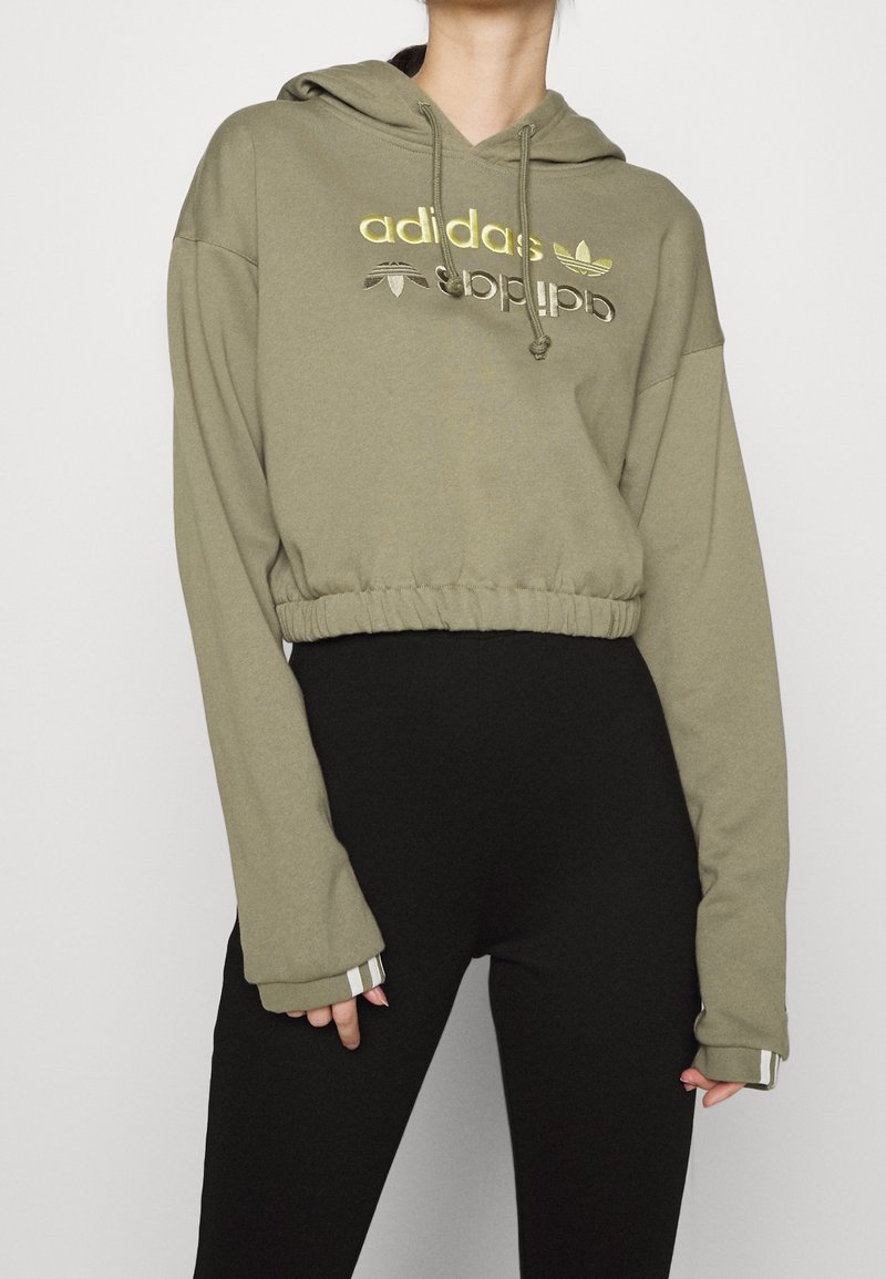 Person wearing olive green cropped hoodie with gold Adidas logo and black high-waisted pants against a plain background.