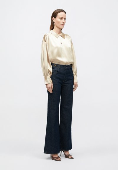 Cămașă bej din satin, cu guler și mâneci bufante, asortată cu blugi evazați din denim închis, având o curea cu accent auriu. Pantofi maro cu degetele goale.