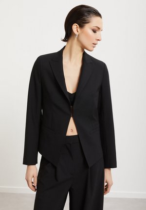Vrouw met haar naar achteren gestyled, die een open zwart colbert draagt over een zwarte top en bijpassende zwarte broek, staand tegen een effen lichte achtergrond.