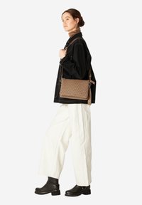 Woven brown leather crossbody bag with a structured rectangular shape, featuring a tassel accent and adjustable strap, worn with white pants.
