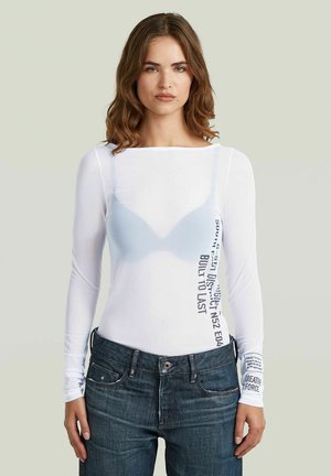 Vrouw met golvend bruin haar die een doorschijnende witte top met lange mouwen draagt over een blauwe beha en donkerblauwe spijkerbroek tegen een effen lichte achtergrond.